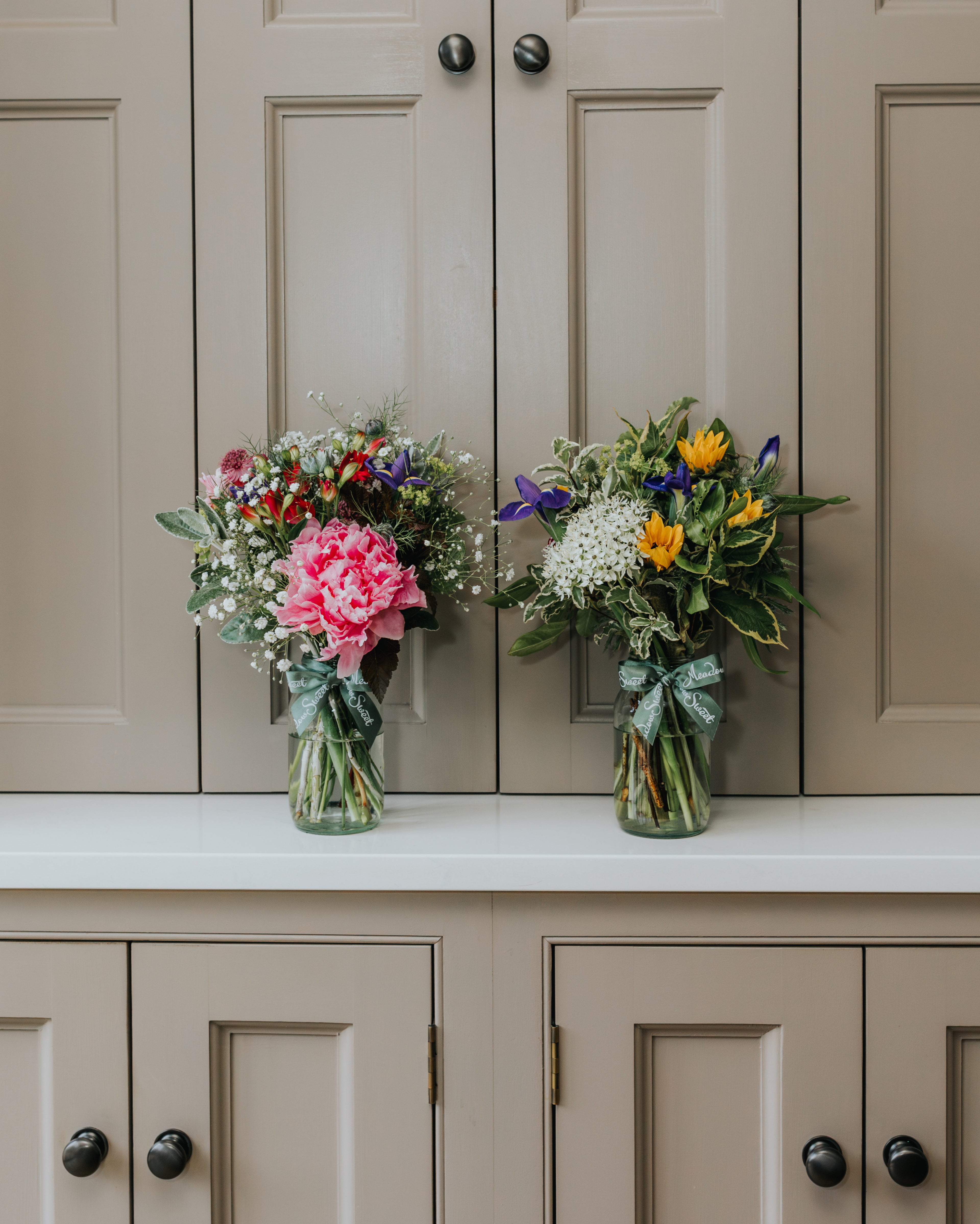 Two MeadowSweet posies of British seasonal flowers in glass jars on kitchen worktop