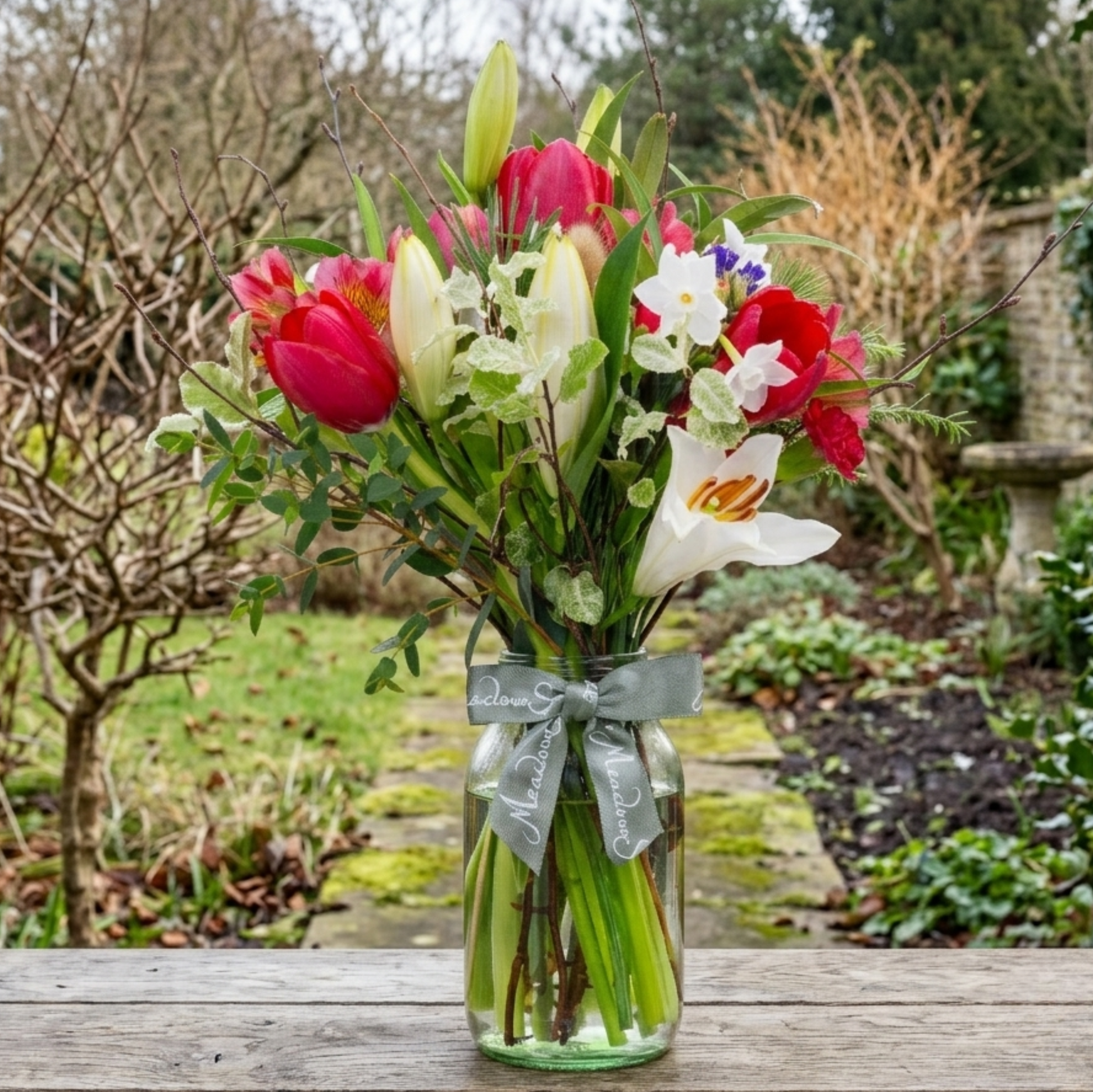 A British flower posy of red tulips, narcissi and lilies in a clear glass jar vase, tied with a ribbon, set on a wooden table in a winter garden.