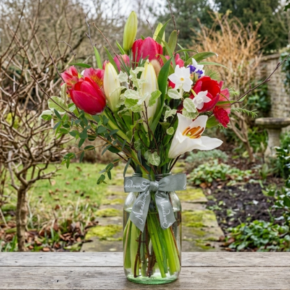 A British flower posy of red tulips, narcissi and lilies in a clear glass jar vase, tied with a ribbon, set on a wooden table in a winter garden.